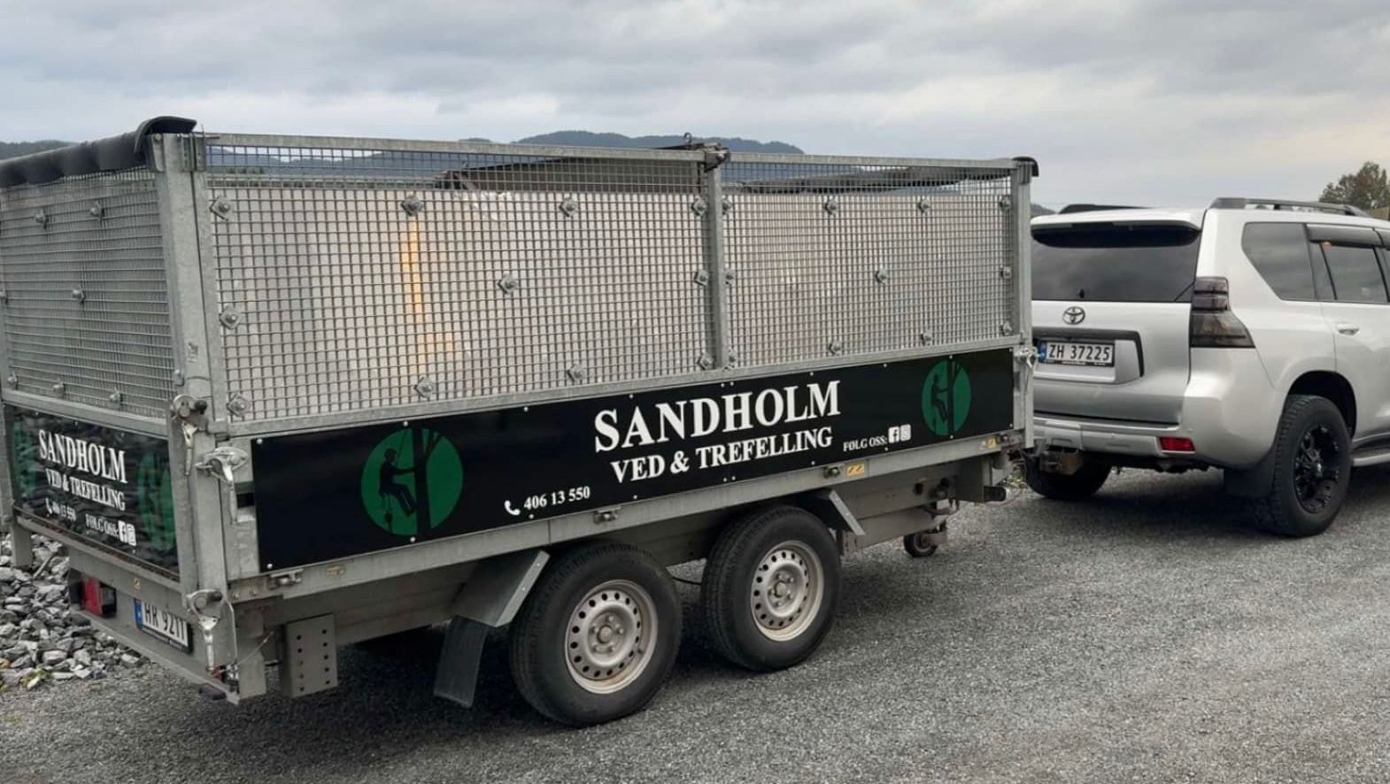 Hvit SUV med tilhenger fra trefellingstjeneste parkert på grusplass under overskyet himmel.