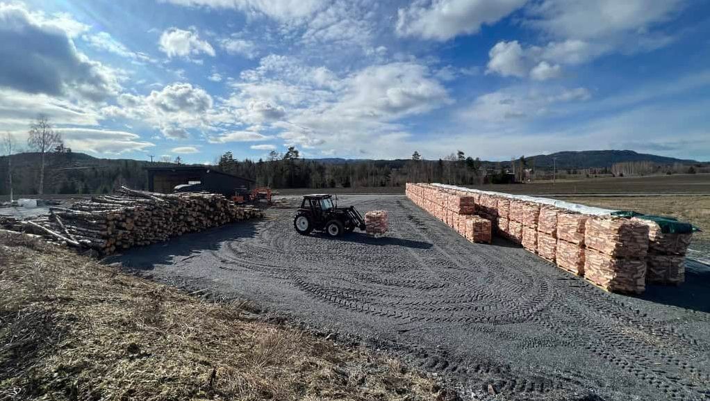 Traktor flytter stabler med trelast på åpen plass med lagret tømmer og materialer rundt.