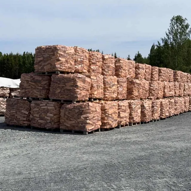 Stablet ved i sekker på paller lagret utendørs på grusplass med skog og blå himmel.