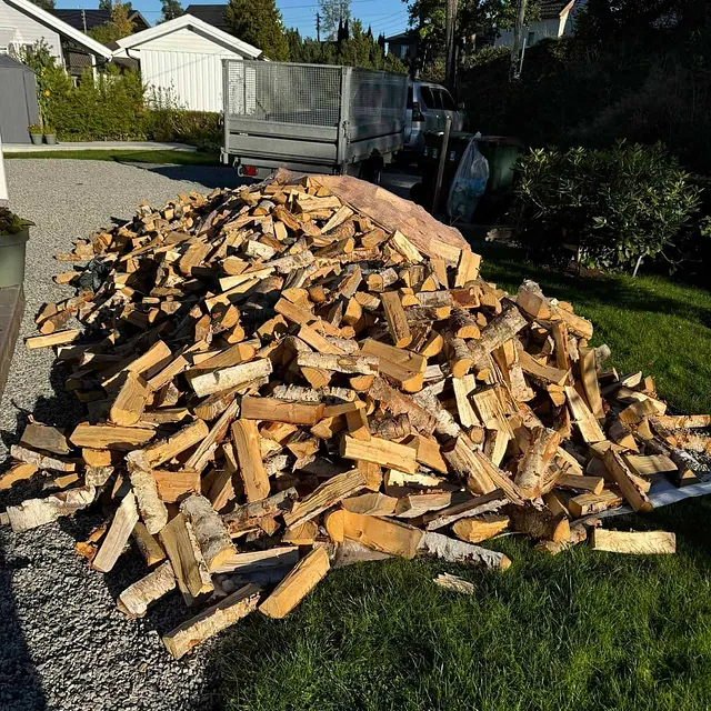 Stor haug med kløyvd ved stablet på gårdsplass ved bolig med tilhenger og hage rundt.