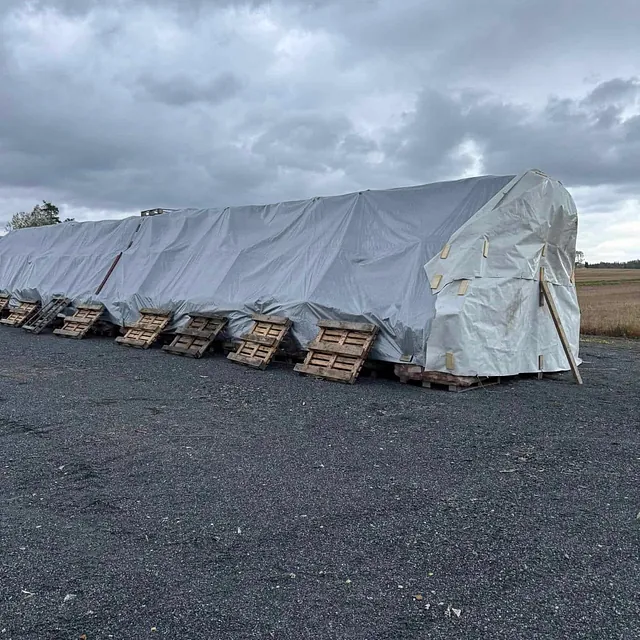 Vedsekker lagret under tett presenning med paller langs siden på grusplass i overskyet vær.