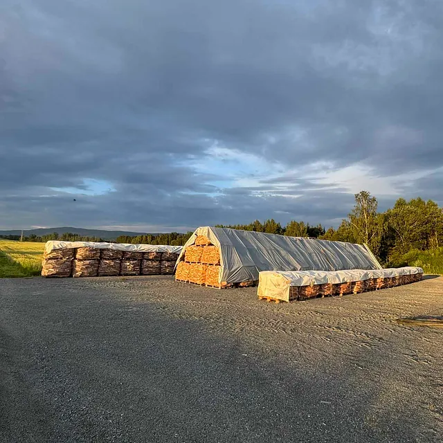 Stablet ved i sekker under presenning på åpen plass med skog og kveldshimmel i bakgrunnen.